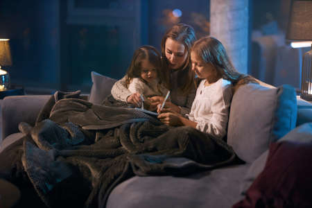 Beautiful caucasian woman reading fairy tale for two little girls while sitting together on cozy sofa. Mother and daughter covered with soft blanket using book before going to sleep.の写真素材