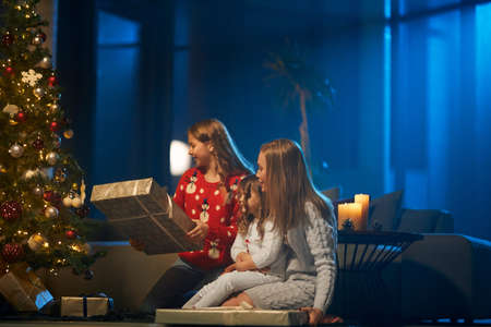 Cheerful mother and her two cute daughters sharing with presents while sitting near christmas tree. Caucasian family celebrating winter holidays together at home.の写真素材