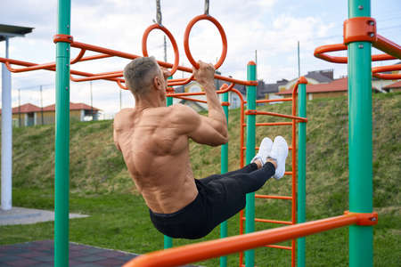 Shirtless caucasian man with muscular body doing abdominal workout on horizontal bar outdoors....