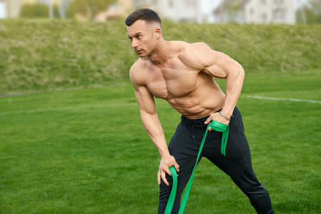 Shirtless caucasian bodybuilder training arms with resistance band on outdoors stadium. Muscular young man enjoying workout on fresh air. Healthy lifestyles.の写真素材