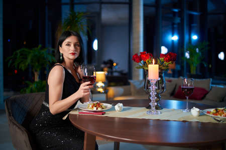 Gorgeous caucasian lady in elegant black dress sitting at table with fresh salad and glass of wine. Romantic dinner at home. Dating concept.の写真素材