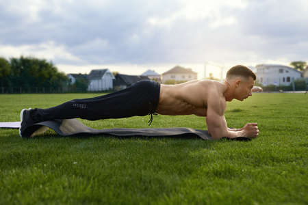 Muscular man with naked torso standing in plank position on yoga mat outdoors. Caucasian bodybuilder doing exercises for abs on grass of stadium.の写真素材