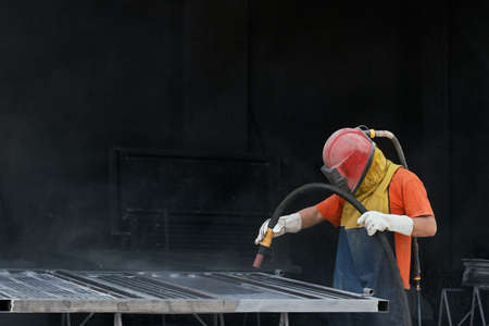 Industrial worker dressed in protective helmet and clothes sandblasting metal details for removing dust and rust. Young man cleaning surface of steel before painting.の写真素材