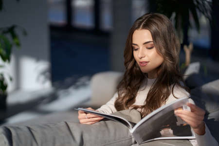 Adorable woman with wavy brown hair reading magazine while sitting on cozy sofa. Attractive lady interesting in fashion news. Relaxation at home.の写真素材