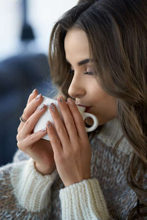 Side view of attractive caucasian woman enjoying tasty coffee while resting at home. Pretty lady with wavy brown hair warming with hot beverage during cold winter days.の写真素材