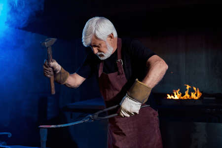 Professional aged blacksmith holding with forceps heated metal and hitting on it with industrial hummer. Bearded man wearing safety apron and gloves. Hard manual work at forge.の写真素材