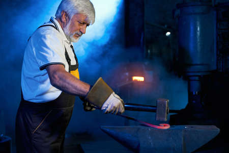 Experienced caucasian blacksmith in protective apron and gloves bending steel on anvil. Mature industrial worker using forceps and hammer at forge.の写真素材