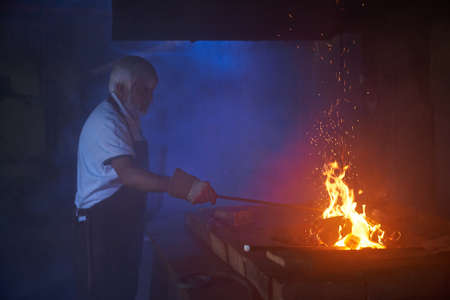 Skillful caucasian blacksmith with grey beard and hair heating steel over burning fire. Mature...