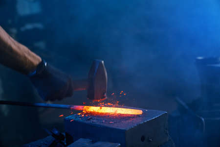 Close up of strong male hands and safety gloves forging molten metal on anvil with hammer. Professional blacksmith working with steel at forge.の写真素材