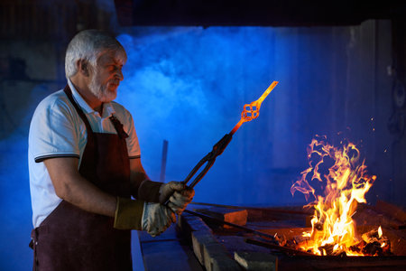 Side view of aged caucasian man in safety apron and gloves heating metal in burning fire at forge....