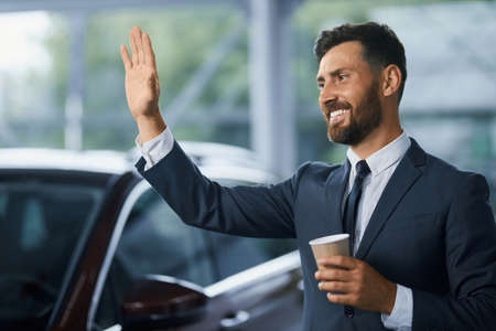 Bearded caucasian man in stylish suit smiling and waving to someone while standing at modern car salon. Businessman with cup of coffee in hand choosing new vehicle.の写真素材
