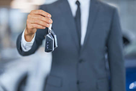 Close up of man in stylish suit holding keys from new modern car while standing at modern showroom. Male customer buying vehicle at salon. Blur background.の写真素材