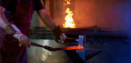 Close up of craftsman in safety gloves processing heated steel on anvil. Strong man using hammer and...