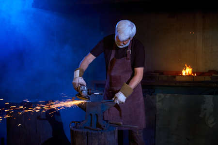 Grey haired man in safety apron and gloves welding metal at workshop with angle grinder. Caucasian...