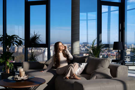 Adorable young woman relaxing on cozy sofa and enjoying sunbeams with closed eyes. Modern apartment...