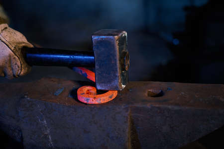 Close up of male hands in safety gloves forging heated steel on anvil using heavy industrial hammer....