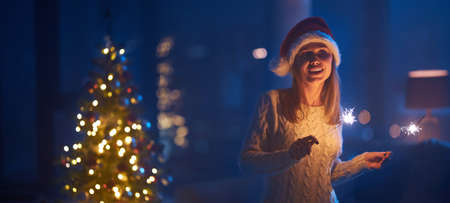Joyful young woman in knitted dress and santa hat celebrating near at home with sparklers in hands. Happy blonde standing near beautiful skinny christmas tree at dark room.の写真素材