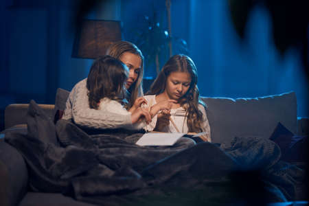 Caring caucasian mother embracing her two little daughters while reading book on couch. Happy family...