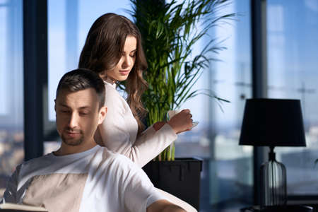 Pleasant woman drinking hot coffee while handsome man sitting on couch and reading book. Young family in casual clothes enjoying relaxation at cozy home.の写真素材