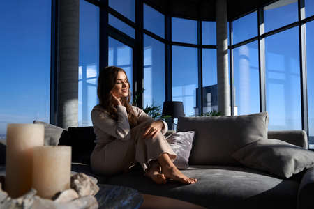 Charming female with dark wavy hair sitting on comfy couch while spending free time at own apartment...