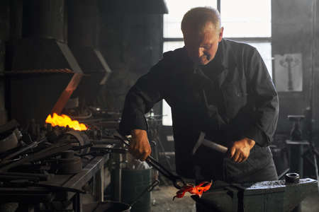 Front view of adult man in nice black uniform working with special tools and creating beautiful decorations for large fence. Concept of process making modern fence in smithy.の写真素材