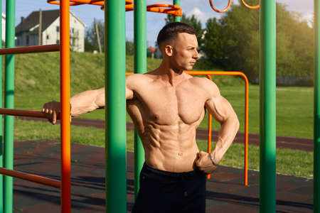 Front view of caucasian male athlete with bare torso looking aside while posting near bars on sports...