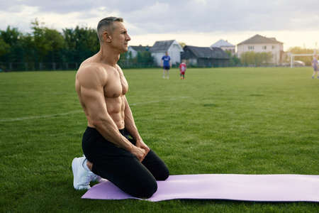 Side view of caucasian man with muscular body shape sitting on yoga mat while resting during...