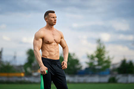 Caucasian man with bare muscular torso standing in outdoors stadium with rubber band in hands. Healthy bodybuilder using sport equipment while training on fresh air.の写真素材