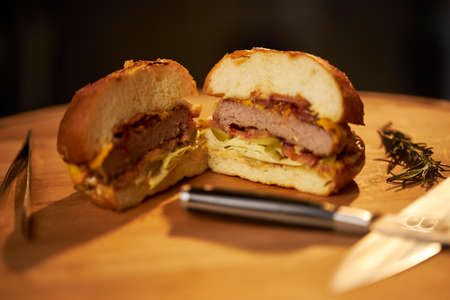 Close up view of grilled beef burger cut in half with knives on cutting board. Juicy homemade cheeseburger with salad, onion and stick of rosemary, isolated on black background. Concept of fast food.の写真素材
