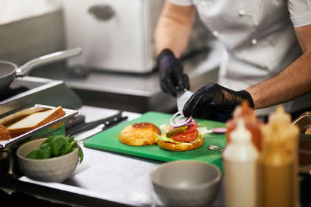Crop view of chefs hands, putting onion rings on delicious hamburger, using spatula in restaurant kitchen. Male hands preparing juicy grilled burger with ingredients on background. Concept of food.の写真素材