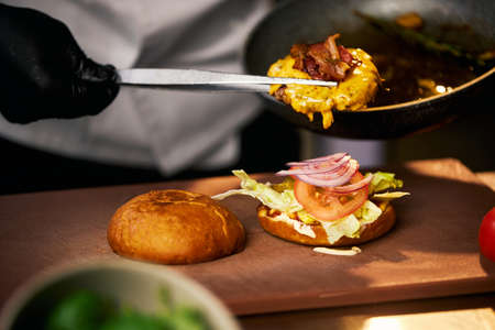Close up view of delicious cheeseburger with red onion, salad and tomato on cutting board. Crop view of chefs hand, putting beef with melted cheese and bacon on bun. Concept of fast food.の写真素材