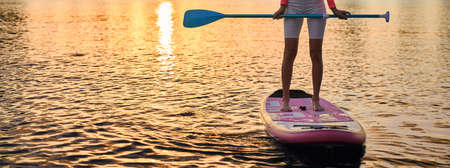 Close up of young woman with slender legs standing on sup board with long paddle in hands. Active female in sportswear spending evening time for water sport.の写真素材