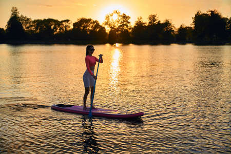 Healthy strong woman in sport outfit surfing on paddle board during summer sunset over lake. Young...