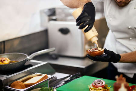 Crop view of chefs hands holding bun, pouring sauce on cheeseburger in restaurant. Male hands in gloves preparing tasty beef burger inside professional kitchen. Concept of food cooking.の写真素材