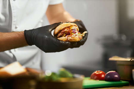 Crop view of head chefs hands in gloves holding tasteful cheeseburger with vegetables. Close up view of mouth-watering grilled burger in restaurant kitchen, with copy space. Concept of fast food.の写真素材