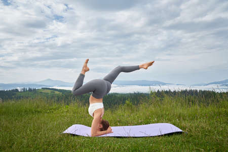 Active young lady wearing sport clothes in headstand position balancing on yoga mat outdoors....