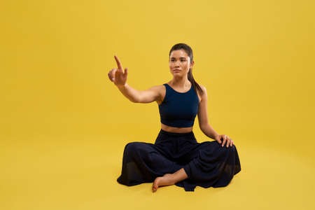 Front view of sporty girl sitting with crossed legs on floor, raising hand, holding finger up,...