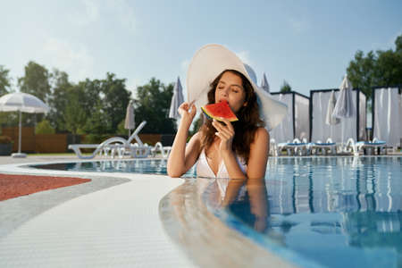 Front view of pretty girl eating watermelon with closed eyes in pool. Seductive young woman standing...