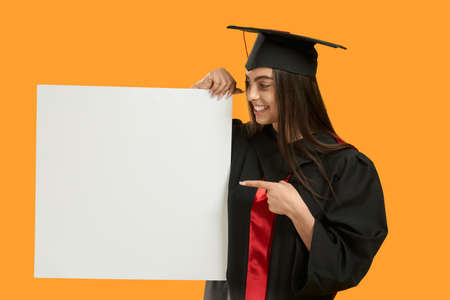 Front view of brunette girl wearing mortarboard and graduate gown, smiling. Pretty young charming female showing, looking down, Isolated on orange mockup studio background.の写真素材