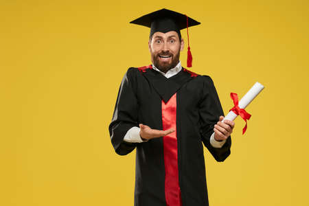 Front view of stdudent standing, holding diploma, surprised. Man with beard wearing graduate gown and mortarboard, looking at camera, smiling. Concept of education and youth.の写真素材