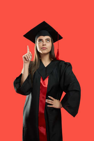 Front view of young female graduating from college, university. Pretty student with bachelor, master degree standing, looking up, thinking. Ioslated on red studeo background.の写真素材