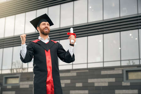 Front view of student wearing mortarboard and graduate gown graduating from college. Brunette boy with beard standing, showing hurray, holding diploma. Concept of high school.の写真素材