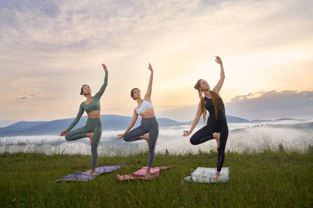 Full length portrait of three young women in sport clothes having yoga practice outdoors. Amazing summer sunset over mountains on background.の写真素材