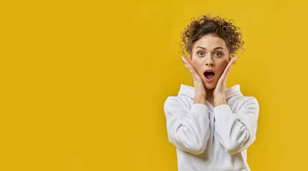 Close up of attractive young woman standing shocked, surprised with open mouth. Pretty slim girl with curly hair touching face by hands, looking at camera. Concept of youth.の写真素材