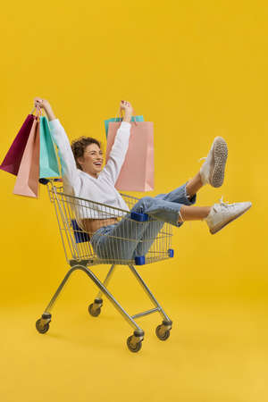 Side view of pretty slim girl sitting in shopping cart. Young blonde woman holding packages, raising hands, looking forward, smiling. Isolated on yellow studio background.の写真素材