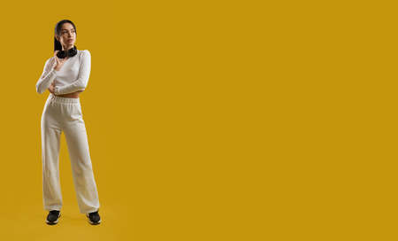 Front view of pretty girl in white sportswear standing with crossed hands, touching hair. Beautiful brunette young female looking aside, thinking. Isolated on yellow background.の写真素材