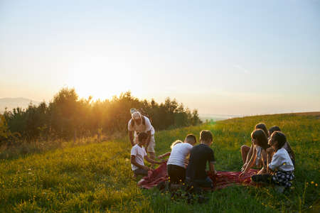 Back panoramic view of group of tourists setting up red tent in hills at sunset. Old man with gray hair explaining, teaching children organizing campsite. Concept of active rest and hiking.の写真素材