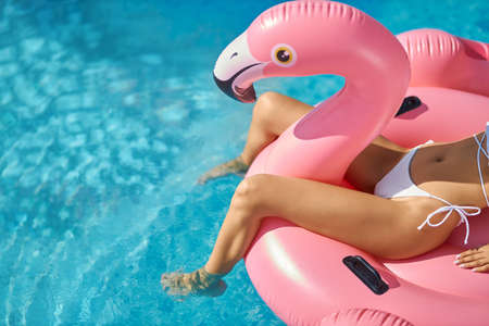 Beautiful tanned female relaxing on coral flamingo bird in swimming pool. Crop view of slim woman in...