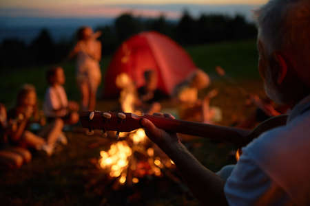 Blurred group of teen friends sitting around campfire, having fun in mountains. Crop view of adult bearded man playing guitar and singing to kids, while camping in evening. Concept of camping.の写真素材