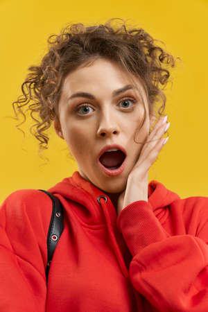 Young woman in red hoodie looking happy and surprised indoor. Portrait view of beautiful girl with wavy hair, dazedly looking at camera, isolated on orange studio background. Concept of emotions.の写真素材
